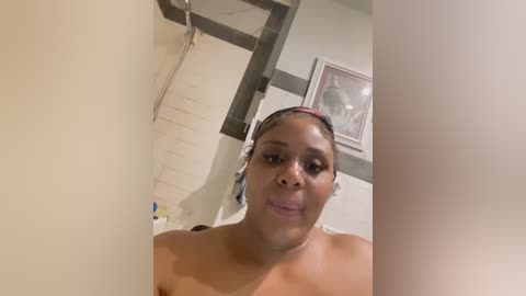 Video of a topless, light-skinned Black woman with braids, wearing headphones, in a shower with beige walls and framed artwork.