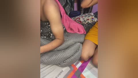 Media: Video of a person with medium skin tone, lying on a bed with a striped blanket, wearing a pink top and a patterned skirt, partially covered by a red blanket.