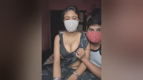 Media: Video of a South Asian woman with medium skin tone, wearing a black patterned dress, mask, and bracelets, sitting on a bed with a young boy in a red mask.