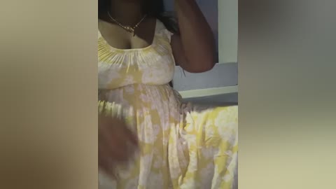 Media: A video of a pregnant woman with medium brown skin, wearing a yellow floral sundress, taking a selfie in a dimly lit room.