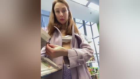 A video of a light-skinned woman with long brown hair, wearing a light pink coat, holding a magazine at a grocery store, with shelves and a large window in the background.