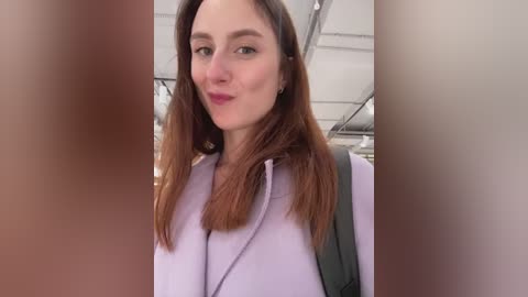 Video of a Caucasian woman with straight, shoulder-length auburn hair, wearing a lavender blouse and a black backpack, smiling softly, standing in a modern, indoor setting with white ceilings and fluorescent lighting.