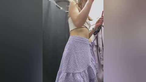 Media: Video of a light-skinned woman with long blonde hair, wearing a white crop top and lavender patterned skirt, taking a selfie in a fitting room with gray walls and a beige garment on the right.