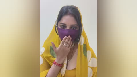 Media: Video of a South Asian woman with medium-brown skin, wearing a yellow sari with white polka dots, purple face mask, and red bangles, covering her mouth, against a plain white background.