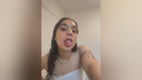 Media: Video of a young woman with medium-brown skin, long black hair, and a tattoo on her right arm. She is sticking out her tongue, wearing a white strapless top, in a minimalist room with cream-colored walls.