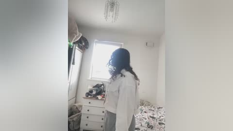 Media: Video of a young woman with curly black hair, wearing a white hoodie and grey skirt, standing in a minimalist, white-walled bedroom with a window, dresser, and bed with patterned bedding.