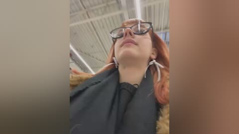 Video of a young woman with fair skin and red hair, wearing black-rimmed glasses and a black shirt, standing in an industrial building with exposed beams and fluorescent lights.