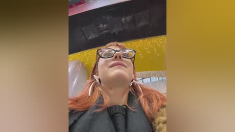 Video of a young woman with red hair and glasses, wearing a black dress, seated at a table with a yellow and silver glitter backdrop, looking upward.