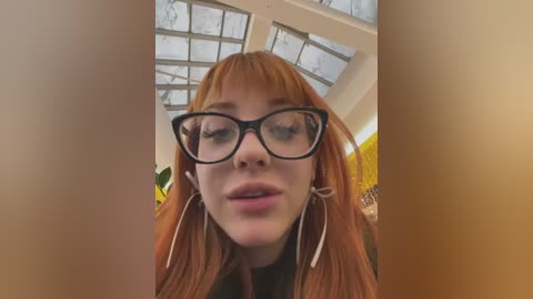 Media: Video of a young woman with long, reddish hair, wearing black-rimmed glasses, white tassel earrings, and a black top. She has a neutral expression, indoors with a skylight and yellow wall visible in the background.