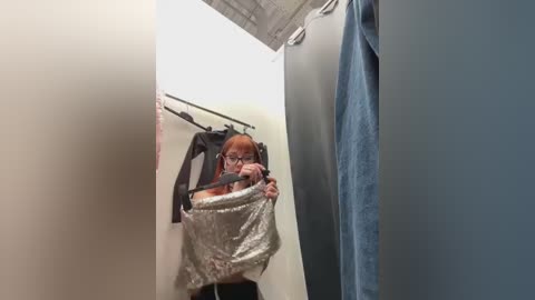 Media: Video of a woman with red hair and glasses, holding a plastic bag, standing in a narrow changing room with beige walls, blue curtain, and a metal rack of clothes.