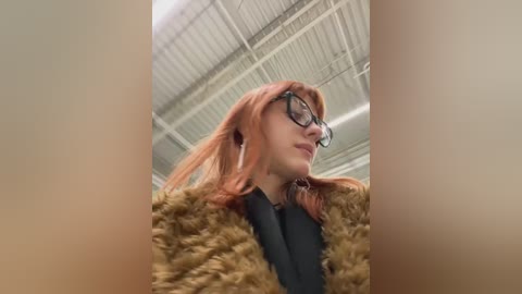 Video of a fair-skinned woman with shoulder-length, reddish-brown hair, wearing large black glasses, a black top, and a fuzzy brown coat, captured from a low angle in a modern, industrial-style room with a high ceiling and white paneling.