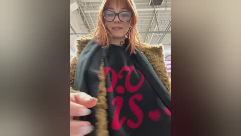 Media: Video of a young woman with straight, shoulder-length orange hair, wearing glasses, a black sweater with pink text, and a furry black jacket. She holds a black object in her hand. Background shows an indoor setting with exposed ceiling beams.