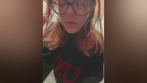 Media: Video of a young woman with pale skin, wearing glasses, orange hair, black top, and large hoop earrings, taken indoors with a blurred background.