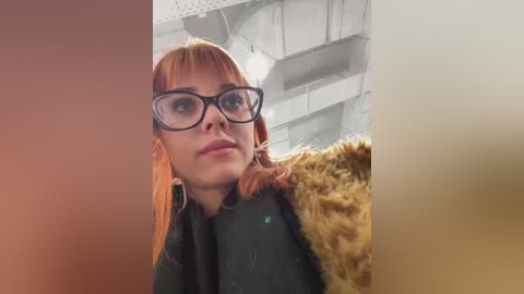 Video of a fair-skinned woman with red hair, wearing black-rimmed glasses and a dark coat, standing in an industrial, white-walled room with exposed beams and pipes.