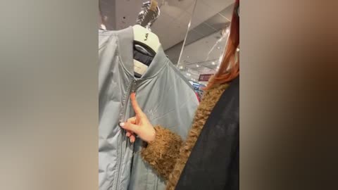 Video of a person wearing a light blue, puffy jacket with a white visor, adjusting the zipper, standing indoors near a mirror with a tan fur-trimmed shoulder bag visible.