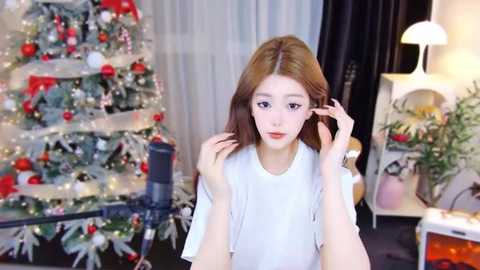 Video of a young woman with light skin, straight brown hair, and a slender physique, wearing a white top. She's indoors, near a decorated Christmas tree and a lamp, adjusting her hair.