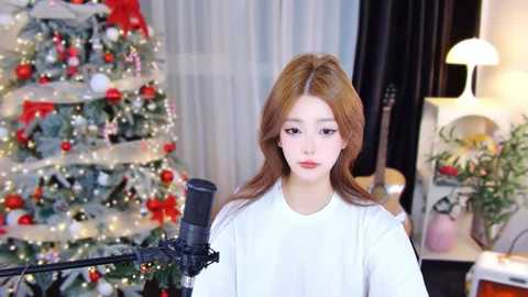 Media: A video of a young woman with fair skin and long, wavy auburn hair, wearing a white top, singing into a microphone in a warmly lit room with a decorated Christmas tree and a guitar.