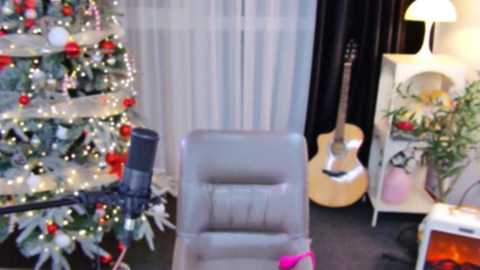 Media: Video of a cozy, festive room featuring a lit Christmas tree adorned with red ornaments, a gray chair, a guitar, and a white lamp on a black shelf.