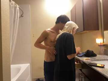 Media: A video depicts a young, shirtless man with a slim build and dark hair, standing in a bathroom, kissing a middle-aged woman with long blonde hair and a dark t-shirt. The background features beige walls, a granite countertop, and a bathtub.