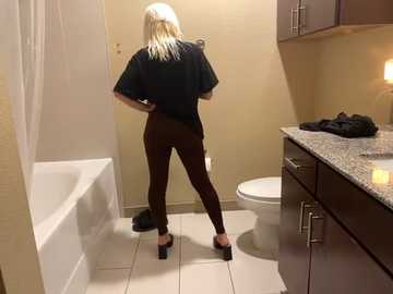 Media: Video of a woman with long platinum blonde hair, wearing a black t-shirt and tight brown leggings, standing in a bathroom, facing a toilet and white bathtub.