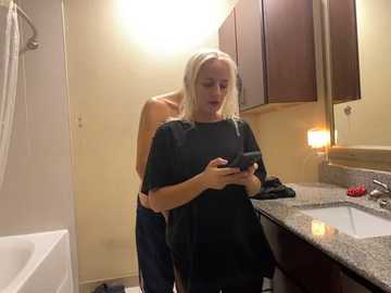 Media: Video of a blonde woman wearing a black cape, texting, in a modern bathroom with beige walls, a granite countertop, wooden cabinets, and a white bathtub.