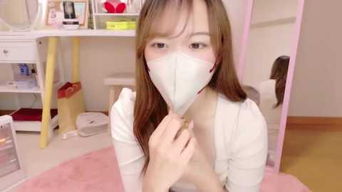 Media: Video of an Asian woman with light skin, wearing a white face mask and a white shirt, kneeling on a pink rug in a tidy, softly lit bedroom with pastel-colored furniture.