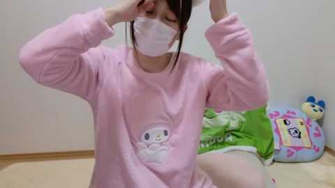 Media: Video of a young Asian woman in a pink hoodie with a cat graphic, wearing a white face mask, adjusting her glasses indoors on a light-colored floor.