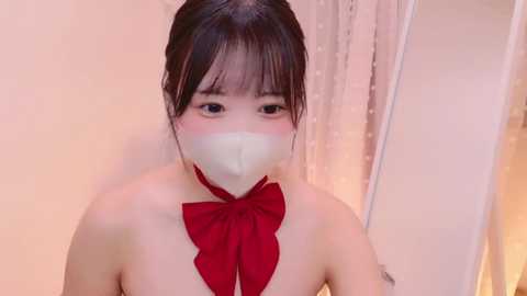 Video of an East Asian woman with light skin, wearing a red bow choker, white surgical mask, and dark hair styled in a bun. She has a calm expression. Background includes a beige curtain and white wall.