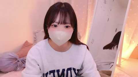 Media: Video of an Asian woman with straight black hair, wearing a white mask, white sweater, and light pink pajamas, sitting in a cozy bedroom with soft lighting, a mirror, and a plush toy.