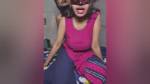 Media: Video of a young woman with dark skin, medium build, and medium-length black hair, wearing a pink patterned dress, sitting on a bed with blue and white striped sheets. She is wearing large, dark sunglasses.