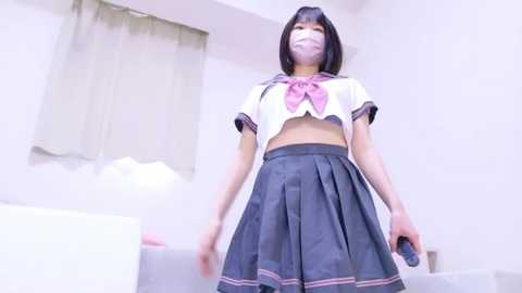 Media: A video of an East Asian woman wearing a schoolgirl outfit, featuring a white crop top with a large pink bow and a pleated gray skirt. She has a surgical mask and a smartphone in her hand. The background shows a white room with a bed and a window.