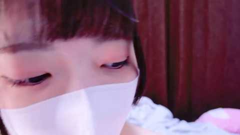 Media: Video of an Asian woman with straight black hair, wearing a white surgical mask, with a blurred wooden background. She has a neutral expression.