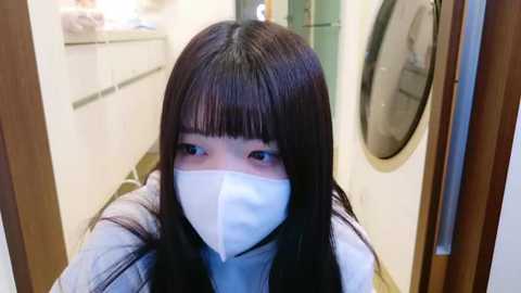 Media: Video of a young Asian woman with long black hair and bangs, wearing a blue surgical mask, standing in a modern laundry room with white cabinets, a washer, and a dryer.