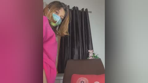 Media: Video of a woman with light brown hair, wearing a pink coat, face mask, and a white shirt, peeking from behind a red curtain, with a brown couch and a pink flower arrangement in the background.