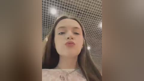 Media: A video of a young woman with long brown hair, fair skin, and closed eyes, puckering her lips. She's wearing a white lace top. The background features a tiled ceiling.