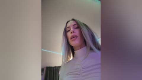 Video of a young woman with long, straight, platinum blonde hair, wearing a white, high-collared top, standing in a dimly lit room with a minimalist decor.