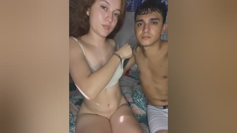 Media: Video of a young, fair-skinned woman with long brown hair, wearing a beige bra, and a man with a light olive complexion, shirtless, sitting on a bed with floral-patterned sheets.