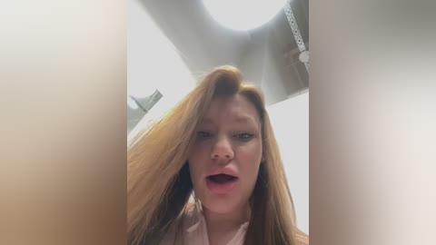 Video of a young woman with long, straight red hair, wearing a light pink top, captured from a low angle, emphasizing her open mouth and surprised expression.