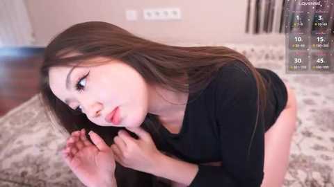 A video of an Asian woman with long brown hair, wearing a black top, leaning forward on a patterned rug in a cozy room.