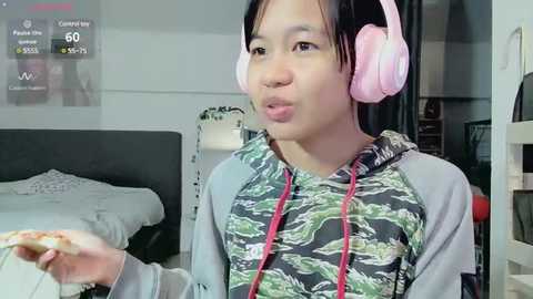 Media: Video of an Asian woman with short black hair wearing pink headphones, gray and green camouflage hoodie, holding a card, in a modern bedroom with a bed, shelves, and a digital clock.