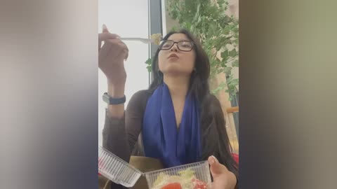 Media: Video of a woman with long black hair, wearing glasses and a blue scarf, holding a lit cigarette and a slice of pizza, indoors near a window with greenery.