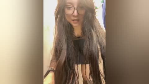 Media: A video of a young woman with long, dark hair and glasses, wearing a black top. She has a light complexion and is indoors with a blurred background.