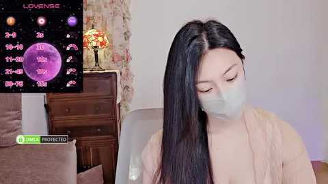 Media: Video of an East Asian woman with long black hair, wearing a white face mask, sitting on a bed. She's dressed in a light-colored robe. Background includes a floral-patterned wall, a wooden dresser, and a vase with flowers.