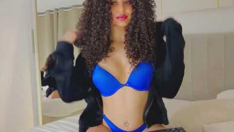 Media: Video of a woman with medium skin tone and curly black hair, wearing a blue bra and panties, black robe, and sitting on a bed in a modern bedroom.