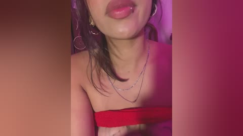 Media: Video of a young woman with light skin, dark hair, and full lips, wearing a red strapless top, silver necklace, and hoop earrings, against a pink background.