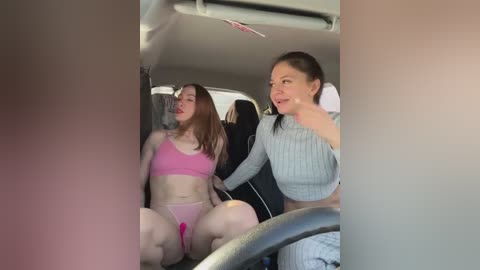 Media: Video of a pink-clad woman with fair skin, seated in a car, holding a black and white striped shirt, while a woman in a grey sweater leans in.