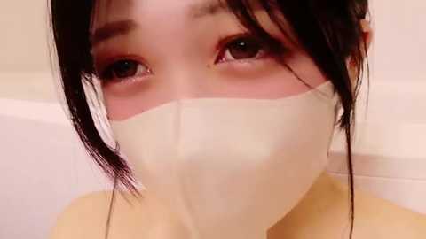 Video of an Asian woman with long black hair and fair skin wearing a white surgical mask, looking directly at the camera. Background is a white bathtub.