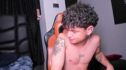 Media: Video of a shirtless, curly-haired, fair-skinned young man with tattoos on his arms, sitting on a black leather chair, in a room with gray walls and a black curtain.