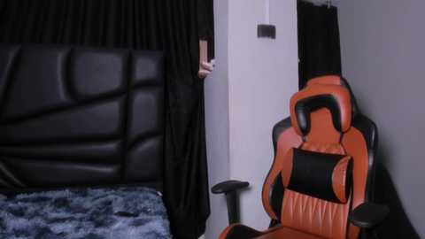 Video of a modern bedroom with a black leather headboard, a blue-patterned blanket, and an orange gaming chair next to a white wall. A person with a black garment stands in the doorway.