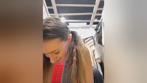 Media: A video of a young woman with long, straight, light brown hair in a ponytail, wearing a sleeveless red top, leaning over to pick something up in a bright, modern, white-tiled room with a skylight.